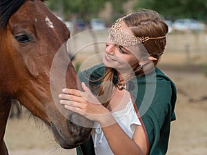 Blonde girl with blue eyes and makeup with elf in the field with a brown horse and a green cape