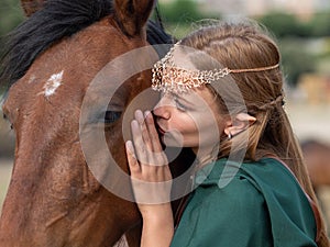 Blonde girl with blue eyes and makeup with elf in the field with a brown horse and a green cape