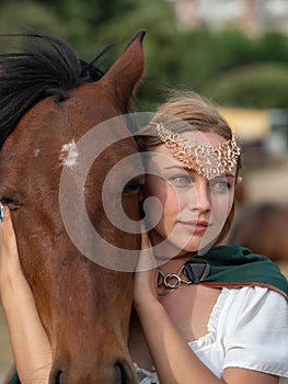 Blonde girl with blue eyes and makeup with elf in the field with a brown horse and a green cape