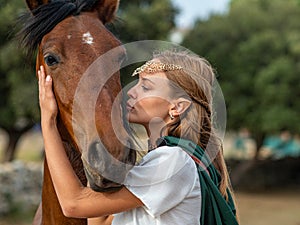 Blonde girl with blue eyes and makeup with elf in the field with a brown horse and a green cape