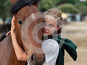 Blonde girl with blue eyes and makeup with elf in the field with a brown horse and a green cape