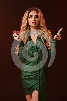 Blonde girl, bright make-up, in green stylish dress and jewelry. Showing two red chips, pointing at something, posing on