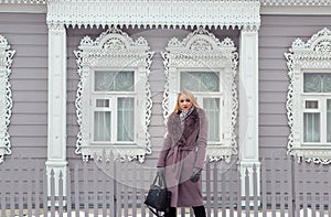 Blonde girl in a brown coat with fur poses on the background of an old Russian wooden house with platbands