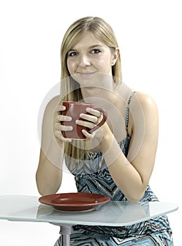 Blonde girl drinking coffee out of a red mug against a white background while sitting at a modern glass desk