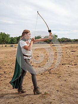 Blonde girl with elf ears poses in the field with a bow and a green cape