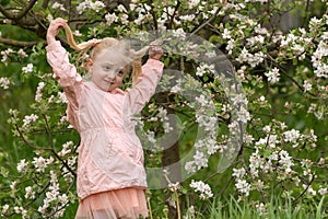 Blonde girl among the flowering trees sits on the grass. Girl with with two tails in next to blooming spring flower tree in spring