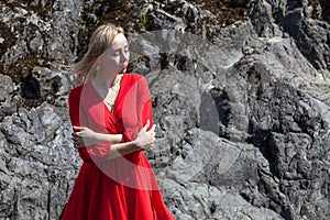 A blonde girl with fluttering hair in the wind in a red dress in an elegant manner stands on the bank of a mountain river with