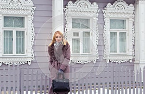 Blonde girl in a gray coat with fur looks into the distance against the background of an old wooden house with platbands