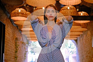 Blonde girl in long blue dress, watch, ring is posing standing under a huge chandeliers in a city cafe. Close-up shot.