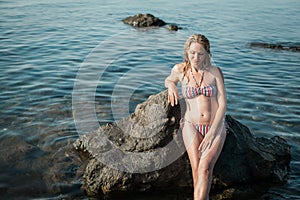 A blonde girl with long hair, in a striped swimsuit, by the ocean, sits by the rocks, in the water and smiles