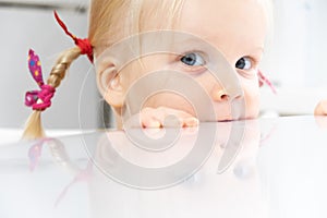 A blonde girl with perky pigtails looks sweetly into the camera and looks out from behind the table on a white