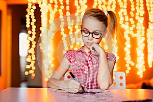 Blonde girl in the pink dress and big black glasses drawing santa claus. Christmas and New Year theme, yellow bokeh