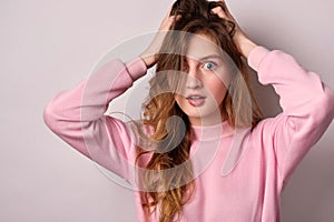 A blonde girl in a pink sweatshirt stands on a white background and looks in horror at the frame ruffling her hair