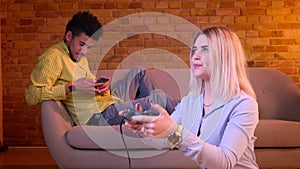 Blonde girl plays videogame on floor attentively with her african boyfriend working with smartphone on background.