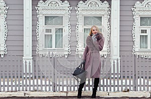 Blonde girl in a purple coat with fur looks away on the background of an old wooden house with platbands