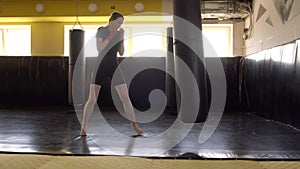 A blonde girl in red bandages beats her hands and feet on a punching bag in the gym. Sports, box, kickboxing, Muay Thai