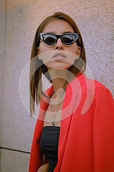 Blonde girl in a red lady-type pantsuit and black top, pendant and sunglasses is posing near a stone wall. Fashion and