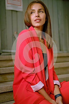 Blonde girl in red lady-type pantsuit and black top, watch, ring, pendant around her neck is posing sitting on a stairs