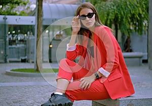 Blonde girl in a red lady-type pantsuit and black top, watch, ring, pendant and sunglasses is posing sitting on a stone
