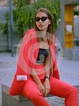Blonde girl in a red lady-type pantsuit and black top, watch, ring, pendant and sunglasses is posing sitting on a stone