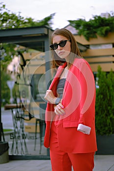 Blonde girl in a red lady-type pantsuit and black top, watch, ring, pendant and sunglasses is walking alone in the city