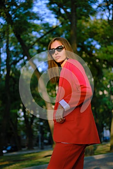 Blonde girl in a red lady-type pantsuit and black top, watch, ring, pendant and sunglasses is walking alone in the city
