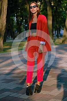 Blonde girl in a red lady-type pantsuit and black top, watch, ring, pendant, sunglasses is walking alone in the city