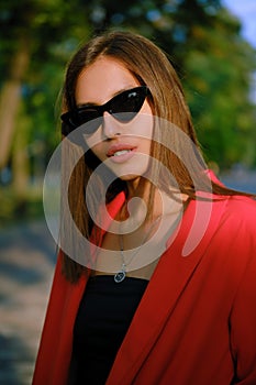Blonde girl in a red lady-type pantsuit and black top, watch, ring, pendant and sunglasses is walking alone in the city