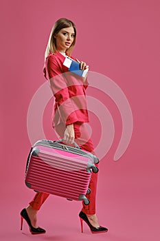 Blonde girl in red pantsuit, high black heels. She is carrying suitcase, holding passport and ticket, pink background