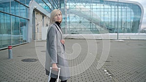 Blonde girl rolls a suitcase near the airport terminal - view from the back