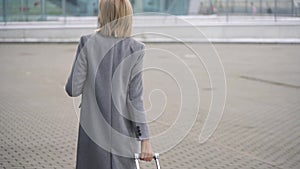 Blonde girl rolls a suitcase near the airport terminal - view from the back