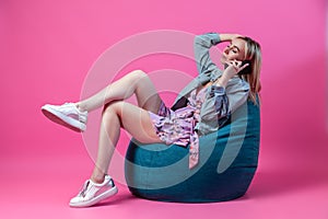 Blonde girl sits in a blue bag chair in a short dress with a neckline and legs cross-legged on a pink background with a phone in