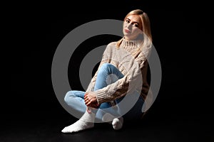 A blonde girl sits on the floor in the studio on a black background in jeans, a sweater and socks, her legs are crossed, she looks