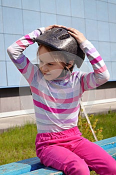 Blonde girl sits in pink jeans on a bench on a bright summer day in a roller helmet in a striped puller with arms raised