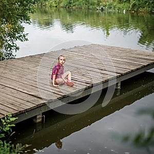 A blonde girl sits on a wooden pier by the river.