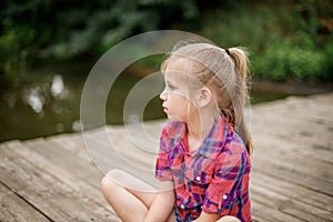 A blonde girl sits on a wooden pier by the river.