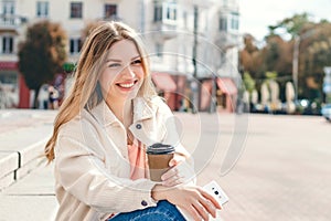 Blonde girl sitting on the steps with a cup of coffee looks away and smiles, copy space