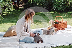 Blonde girl sitting on white blanket in garden and having picnic at sunny day with cute puppies.