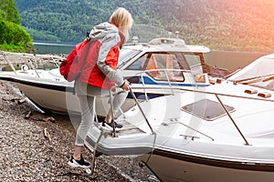 Blonde girl in sportswear and with a red rucksack on her back climbs a boat on the background of the mountain Teletskoye Lake for
