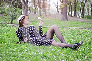 Blonde girl in a straw hat with a dandelion in his hand lies  on the grass in the spring garden.