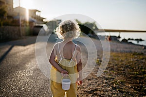 Blonde girl in summer outfit on walk, holding small plastic bucket, going to searchseashells on beach.