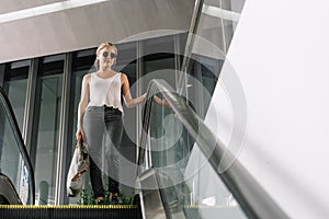 Blonde girl with sunglasses and a grey bag in her hand walking down an escalator