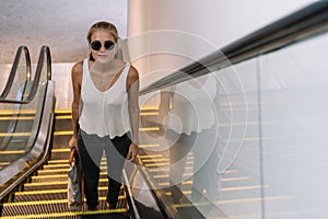 Blonde girl with sunglasses and a handbag climbing an escalator