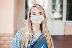 blonde girl wearing a medical mask and standing against the background of a university, college building with folders