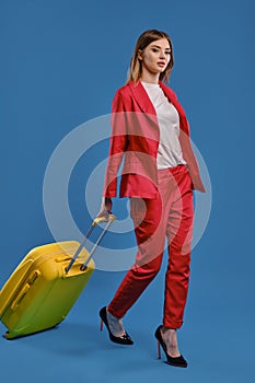 Blonde girl in white blouse, red pantsuit and high black heels. She carrying yellow suitcase by handle while walking on