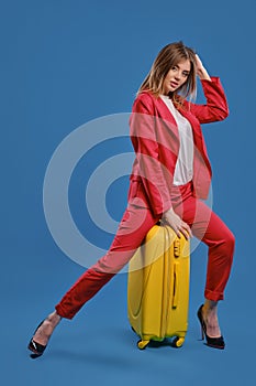 Blonde girl in white blouse, red pantsuit, high black heels. She touching hair, sitting on yellow suitcase, posing on