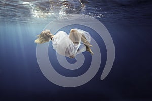 Blonde girl wrapped in fine white cloth, sank in blue deep water of ocean, against dark sea background.