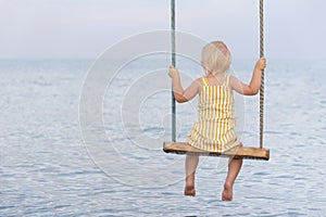Blonde girl in yellow dress sitting on a rope swing over water. Swing on the sea