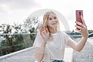 Blonde hair happy young woman posing outdoors in park take a selfie by mobile phone while standing in the park.