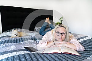 Blonde hair woman sitting on cozy bed with dog, holding open book and reading. Lying woman relaxing on sofa at home